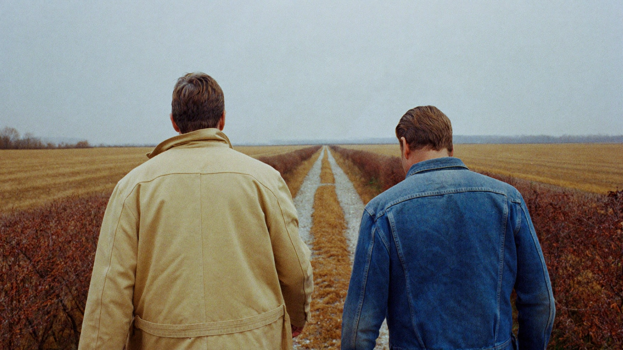 Two men in a chore coat and denim jacket walking a gravel farm track between autumn fields